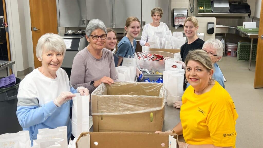 a group of smiling female volunteers lined up on each side of tables with boxes of food between them